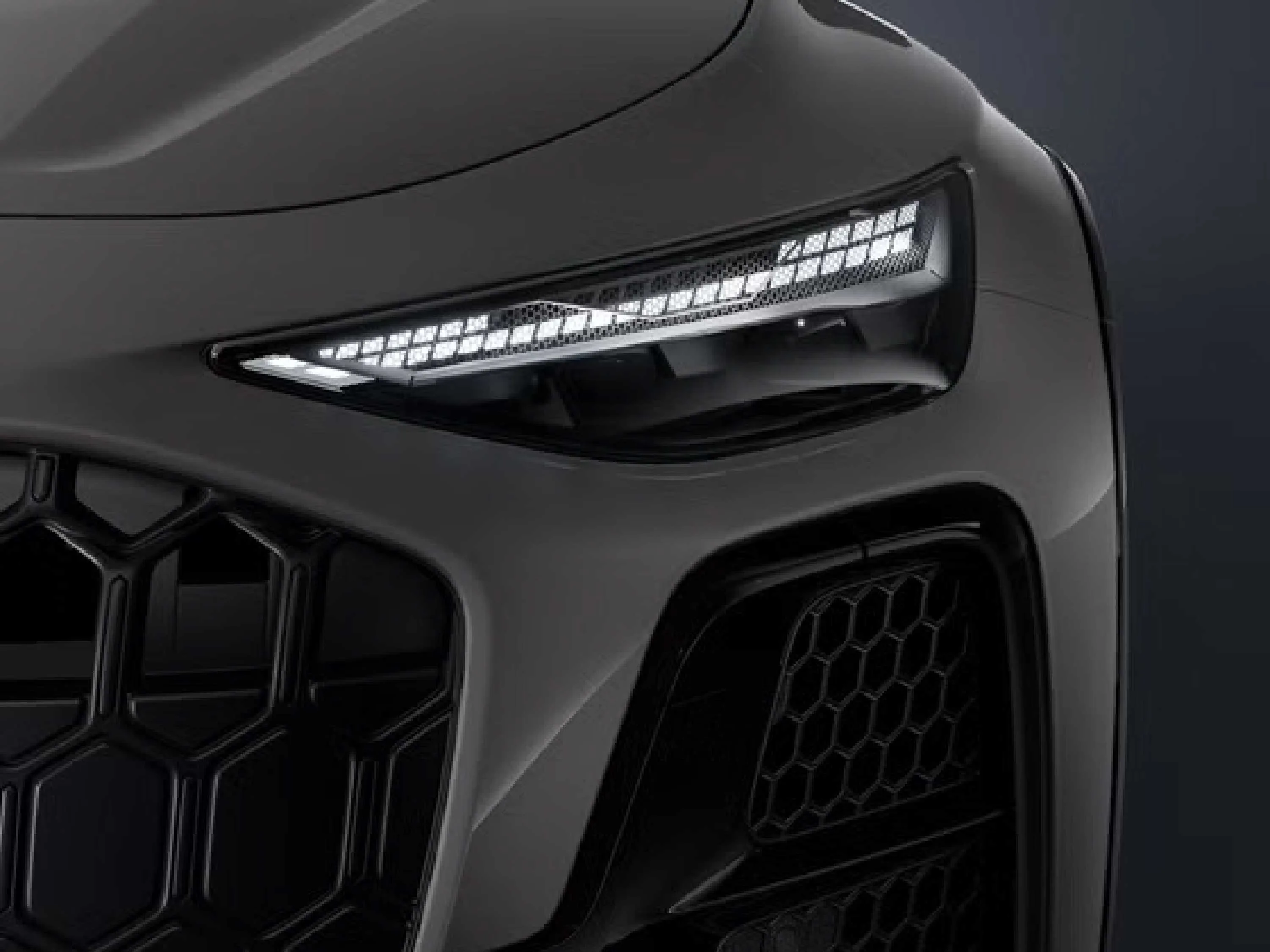 26-073 website galerie 1 Detailaufnahme der schmalen, futuristischen Frontscheinwerfer eines grauen Audi mit leuchtendem, segmentiertem LED-Tagfahrlicht. Ein Ausschnitt des dunklen, wabenförmigen Kühlergrills und der Frontschürze ist im Fokus zu sehen.