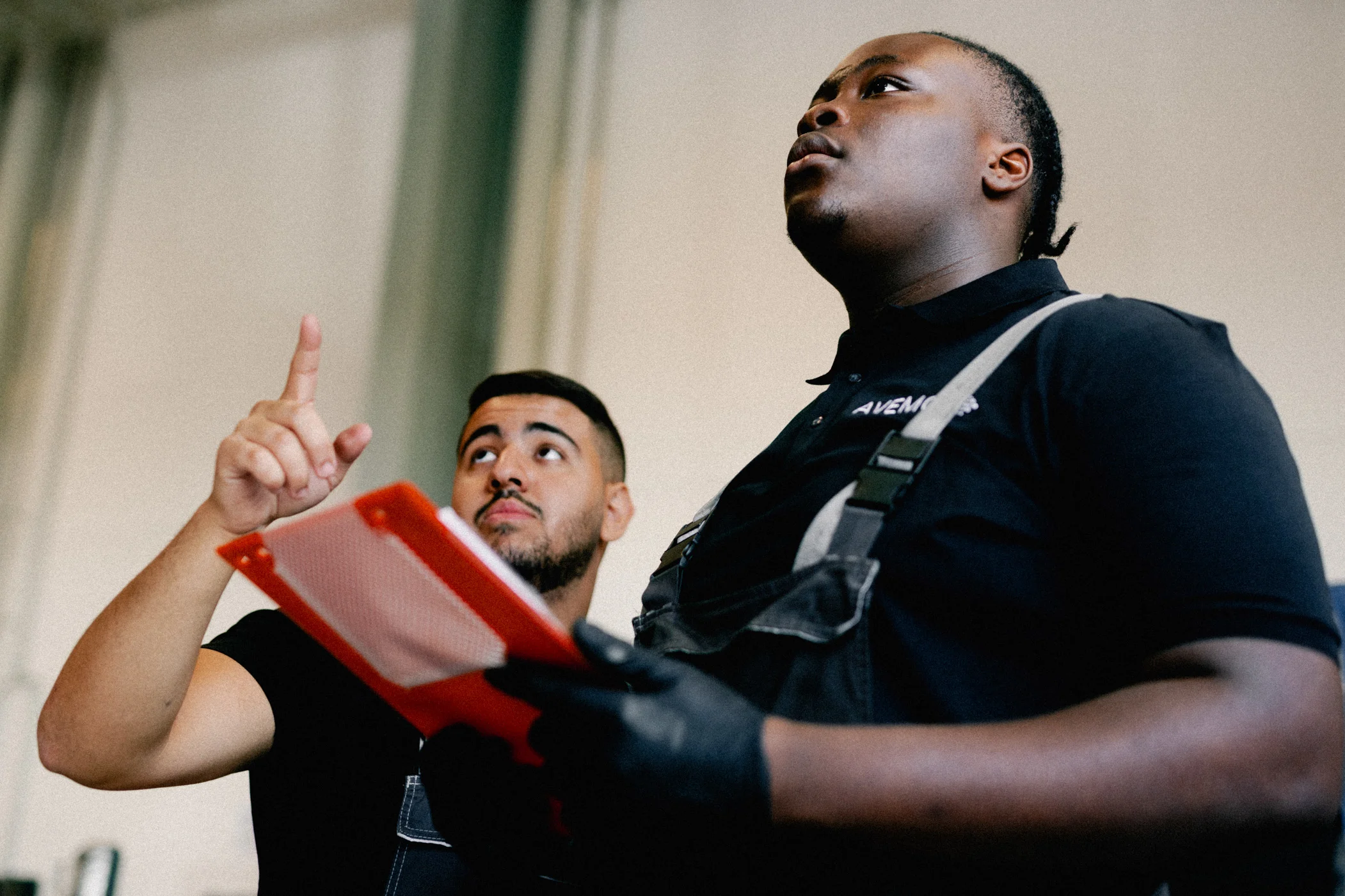 Zwei Männer in Overalls und AVEMO Polo-Hemden bei der Arbeit in einer Werkstatt. Der eine hält mit beiden Händen Unterlagen zu einem Kundenfahrzeug in der Hand. Der andere deutet mit dem Finger auf ein Bauteil des Fahrzeuges.