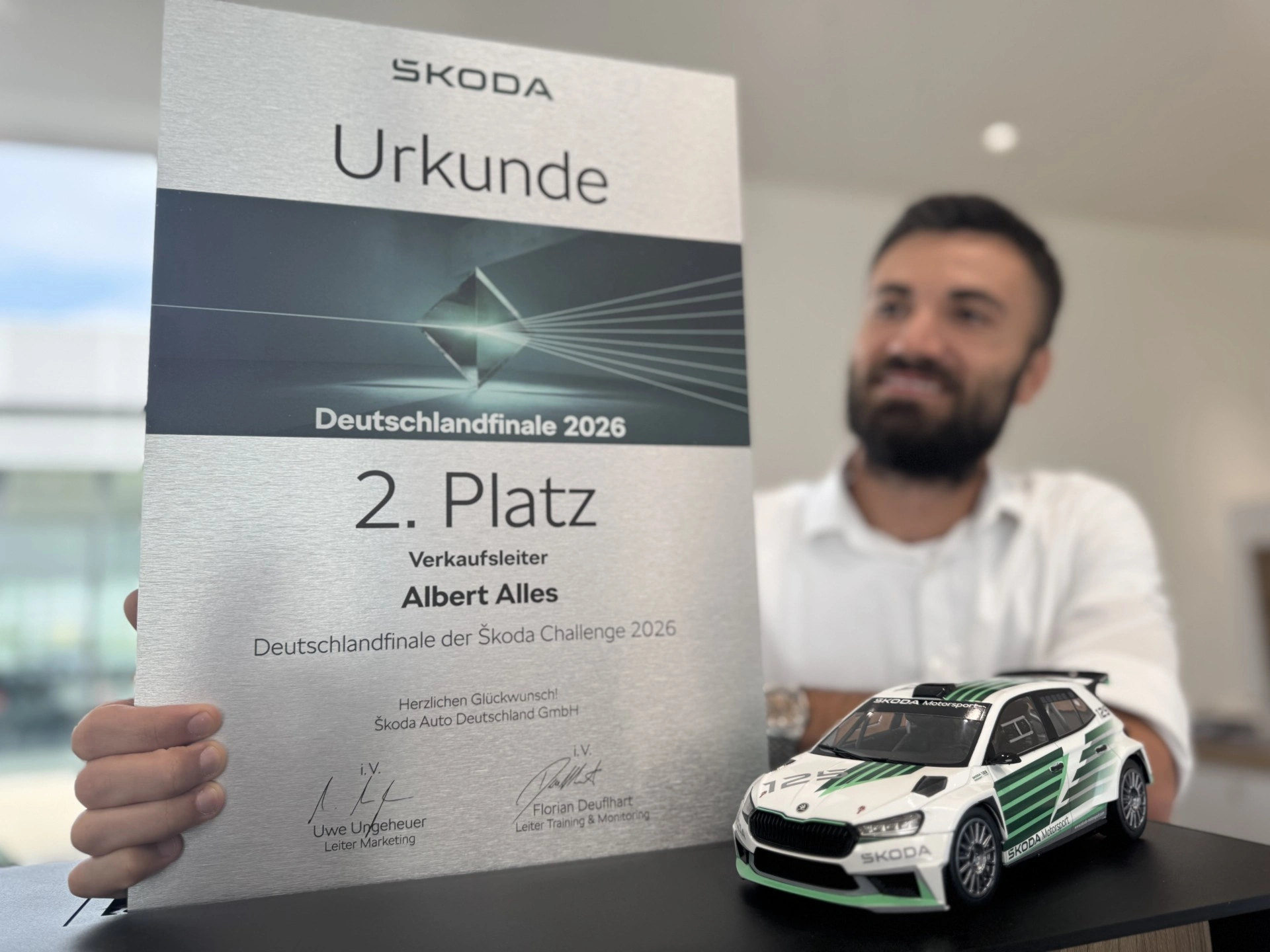 Eine Urkunde der Škoda Challenge 2026 für den 2. Platz im Deutschlandfinale der Verkaufsleiter, gehalten von Albert Alles. Im Vordergrund steht ein Škoda-Motorsport-Modellauto.