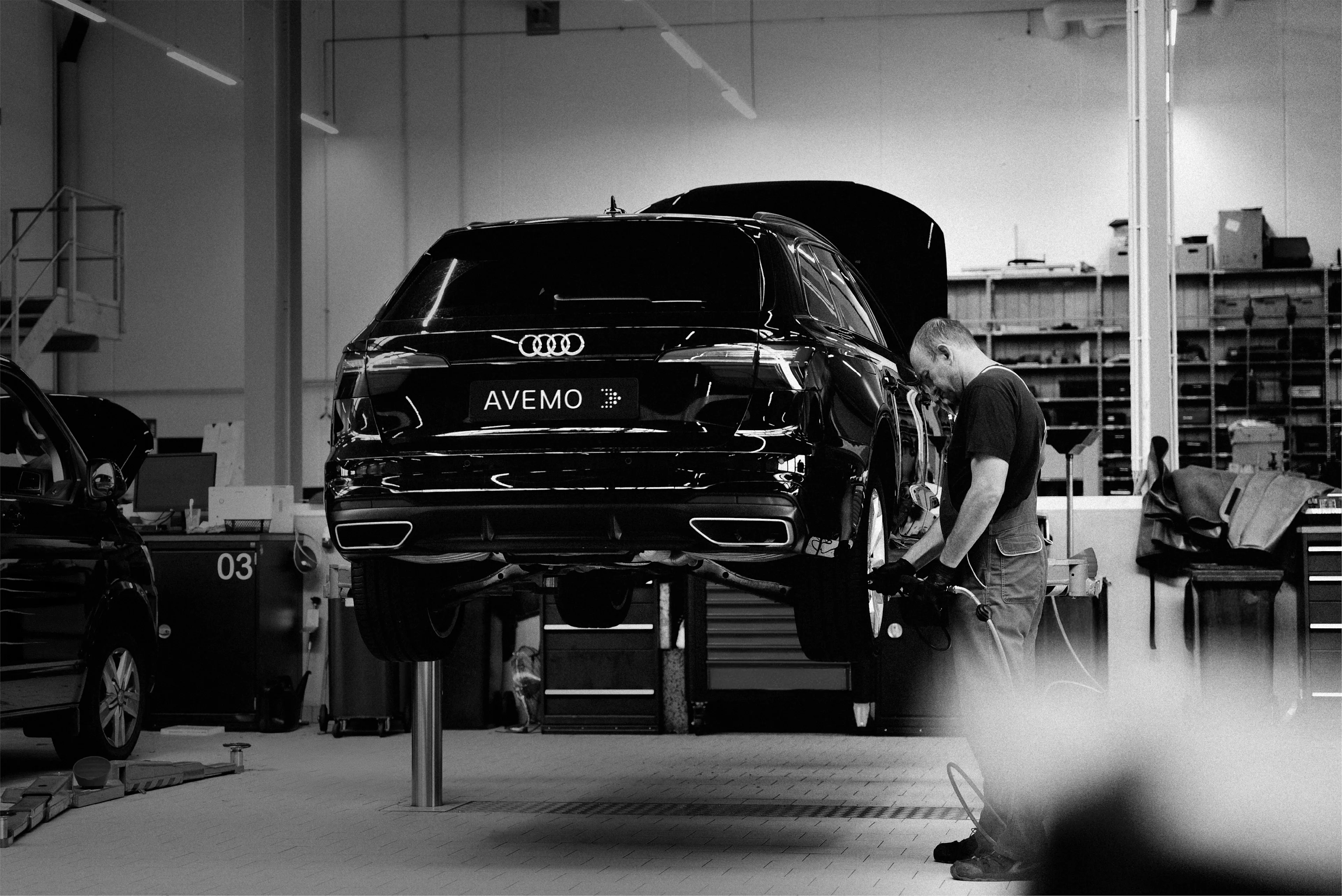 Schwarz-weiß-Fotografie einer Werkstatt: Ein Mechaniker wechselt das Rad an einem schwarzen Audi, der auf einer Hebebühne steht. Das Fahrzeug trägt ein AVEMO-Kennzeichen. Im Hintergrund ist die professionelle Werkstatteinrichtung erkennbar.