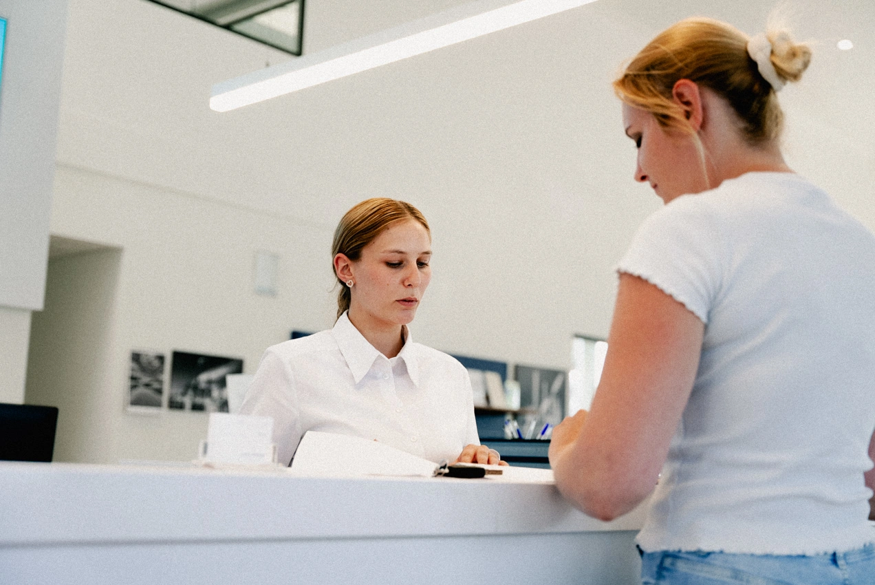 Information Konversation w,w Eine Service-Mitarbeiterin und eine Kundin stehen zusammen am Empfangstresen des Autohauses. Beide unterhalten sich miteinander über Dokumente, die auf dem Tresen vor ihnen liegen.