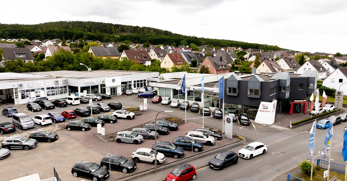 Eine Aufnahme des Volkswagen Autohauses in Lichtenfels, die das Gebäude, den Parkplatz und Fahrzeuge vor dem Gebäude, sowie die Grünewaldstraße und die Einfahrt zeigt.