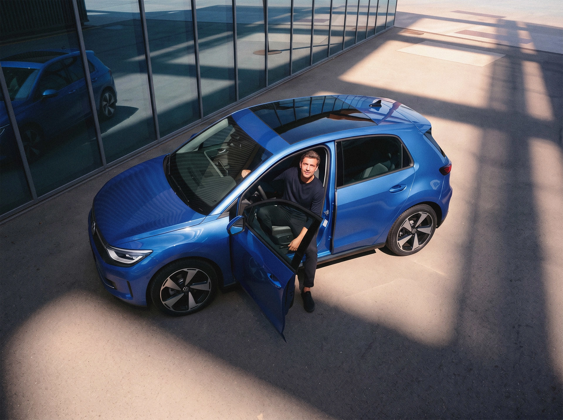 JobMobility – 3 Vogelperspektive auf einen blauen Volkswagen vor einem Bürogebäude: Ein Mann sitzt auf dem Fahrersitz und schaut bei offener Tür nach oben. Ein passendes Motiv für die flexible JobMobility App.