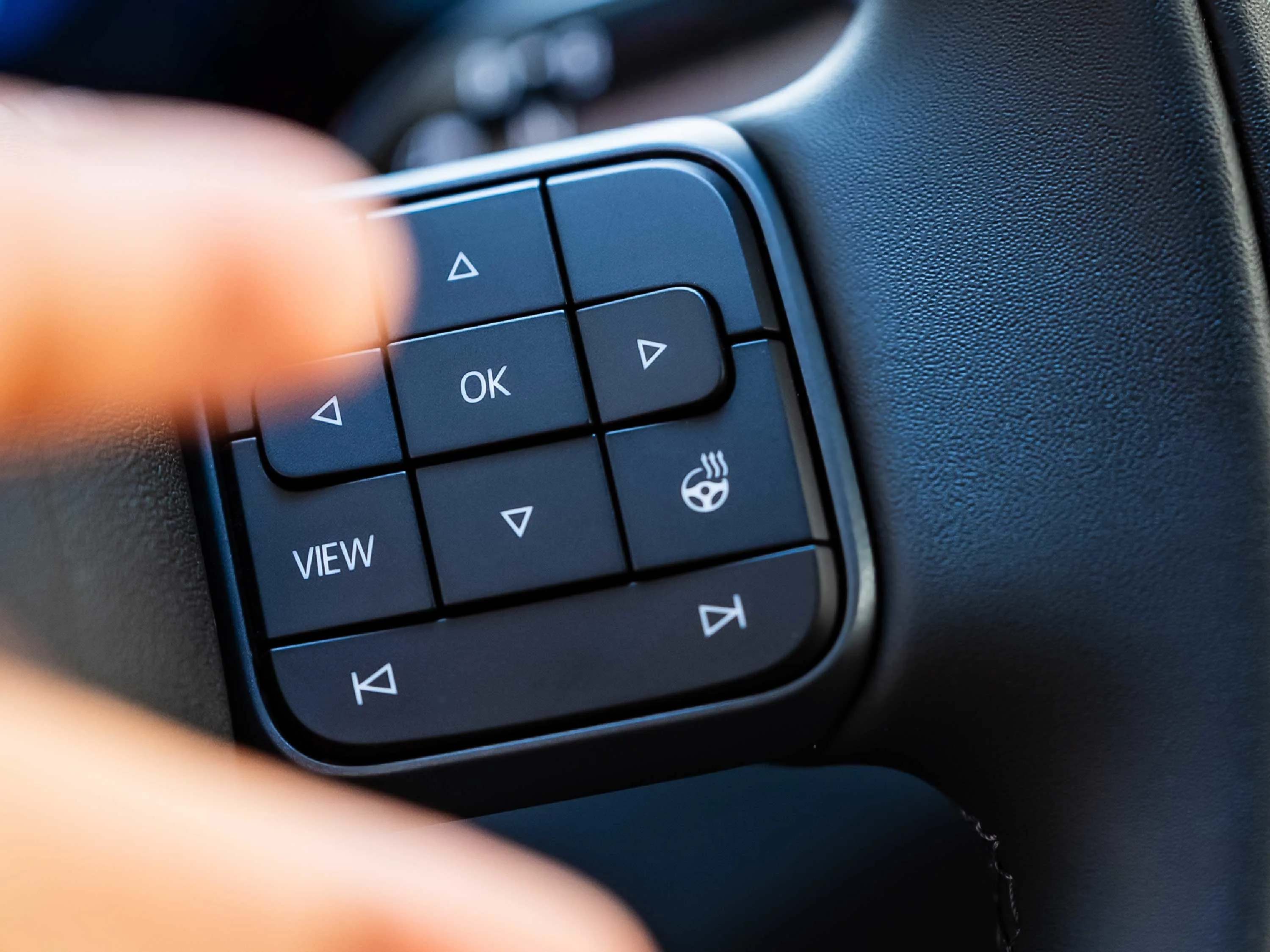 Nahaufnahme der Bedienelemente am Multifunktionslenkrad eines VW ID.3. Zu sehen sind Tasten für die Menüsteuerung (OK, Pfeile, View) sowie das Symbol für die Lenkradheizung. Ein Finger ist unscharf im Vordergrund bei der Bedienung zu erkennen.