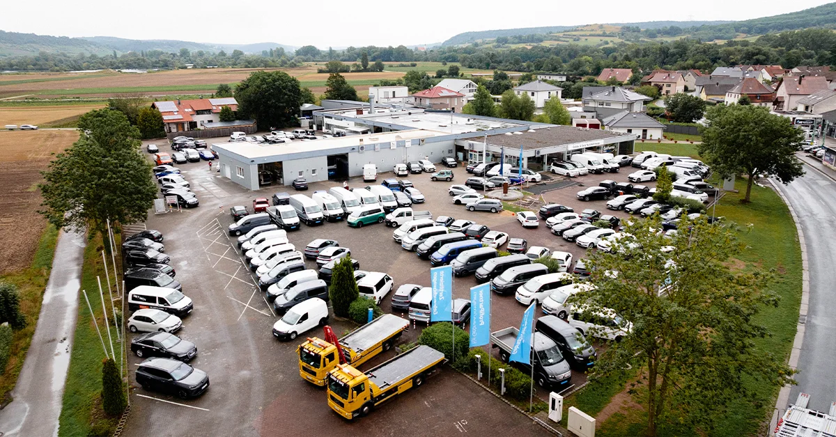 Eine Aufnahme des Volkswagen Nutzfahrzeuge Autohauses in Sand am Main, die das Gebäude, den Parkplatz und Fahrzeuge vor dem Gebäude, sowie die Einfahrt zeigt.
