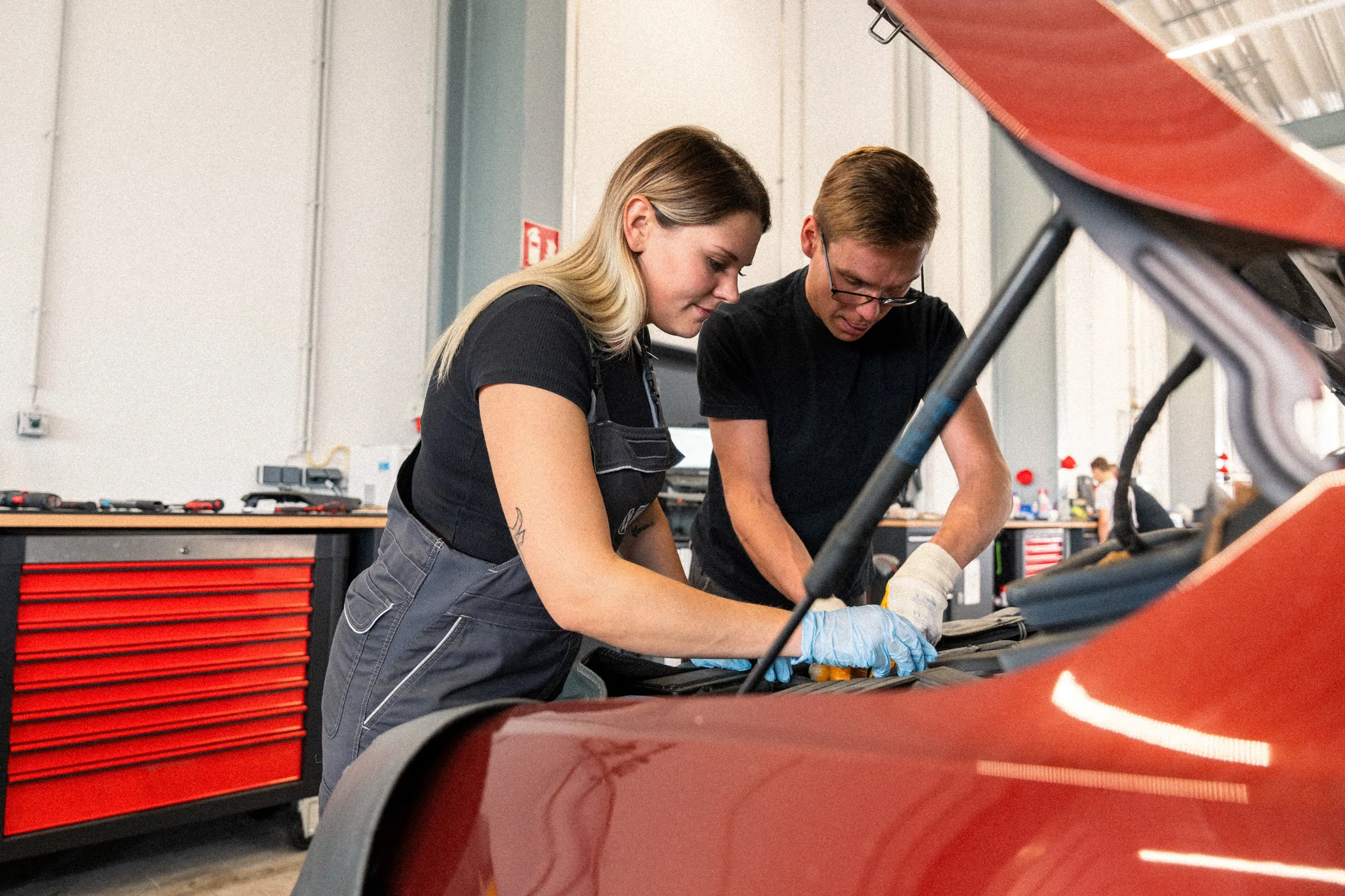 Eine Frau und ein Mann in Arbeitskleidung stehen an der geöffneten Motorhaube eines Auto und führen Service-Arbeiten durch.