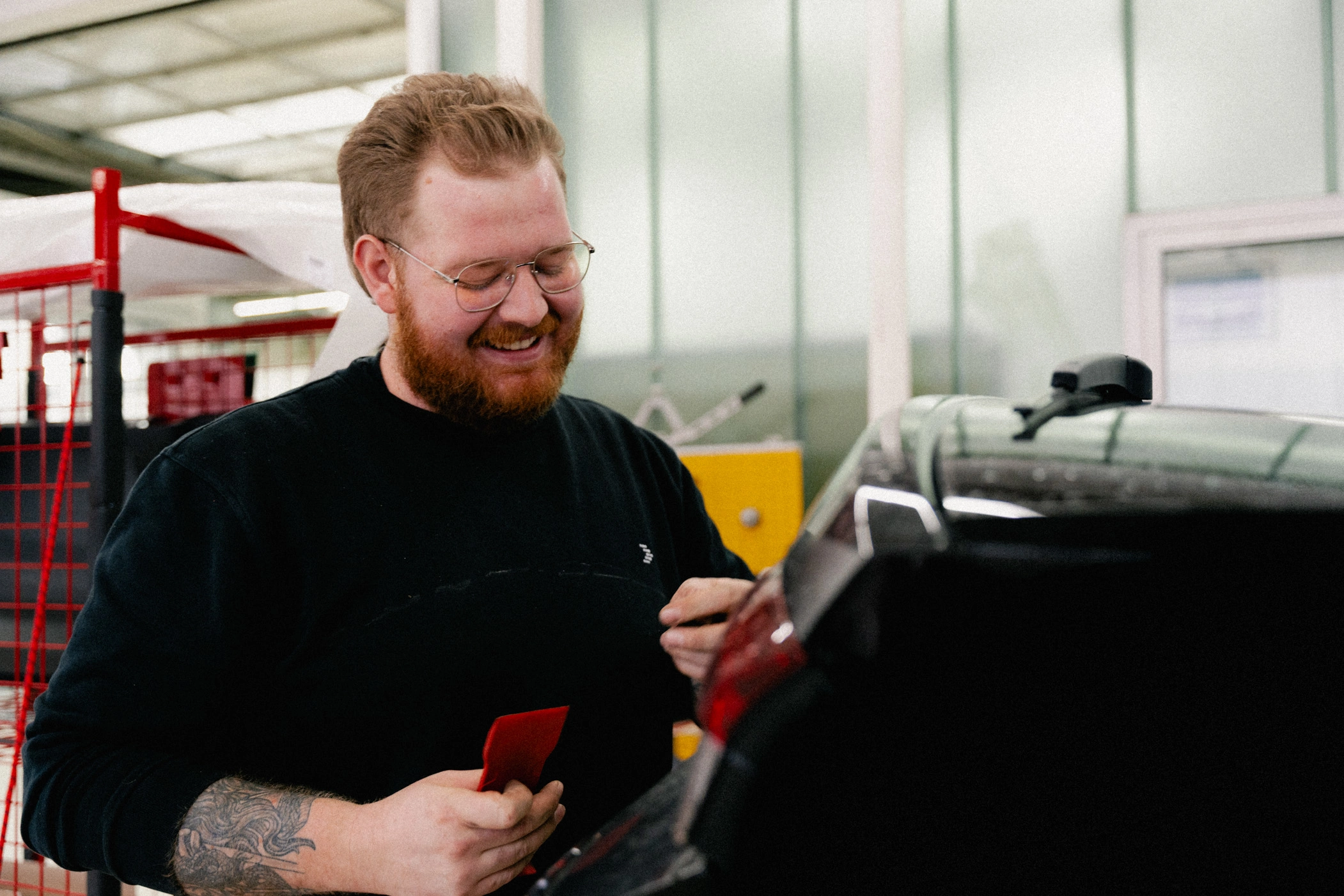 Ein Mitarbeiter steht vor der Heckklappe eines Autos und führt Reparaturen durch. In seiner rechten Hand hält er ein Werkzeug. Außerdem lächelt er und ist bekleidet mit einem schwarzen AVEMO Pullover.