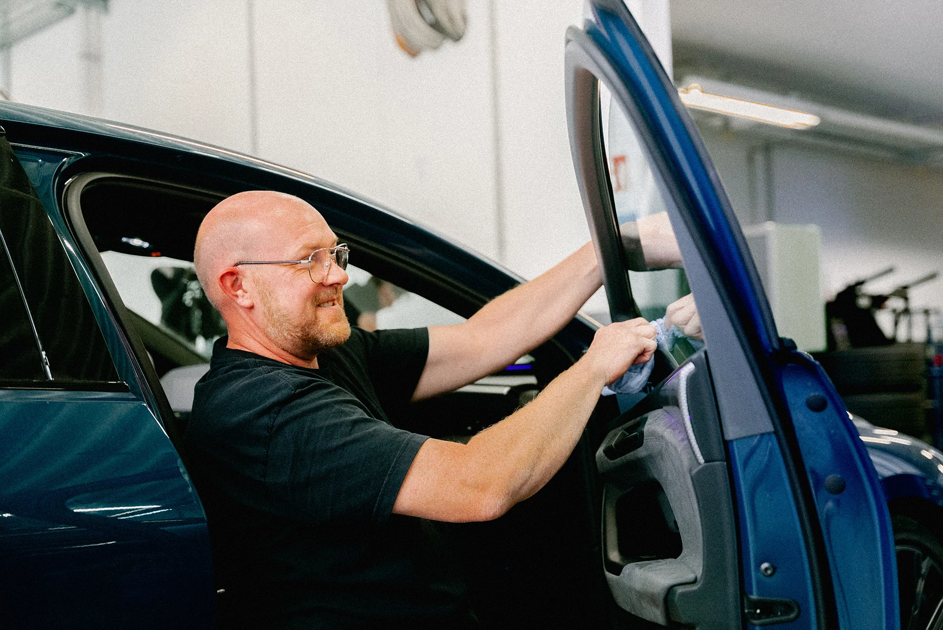 Ein Mann mit Brille steht zwischen der offenen Beifahrertür und dem Fahrzeug und reinigt mit einem Tuch die Beifahrertür-Glasscheibe. Im Hintergrund ist eine helle Werkstatt zu sehen.