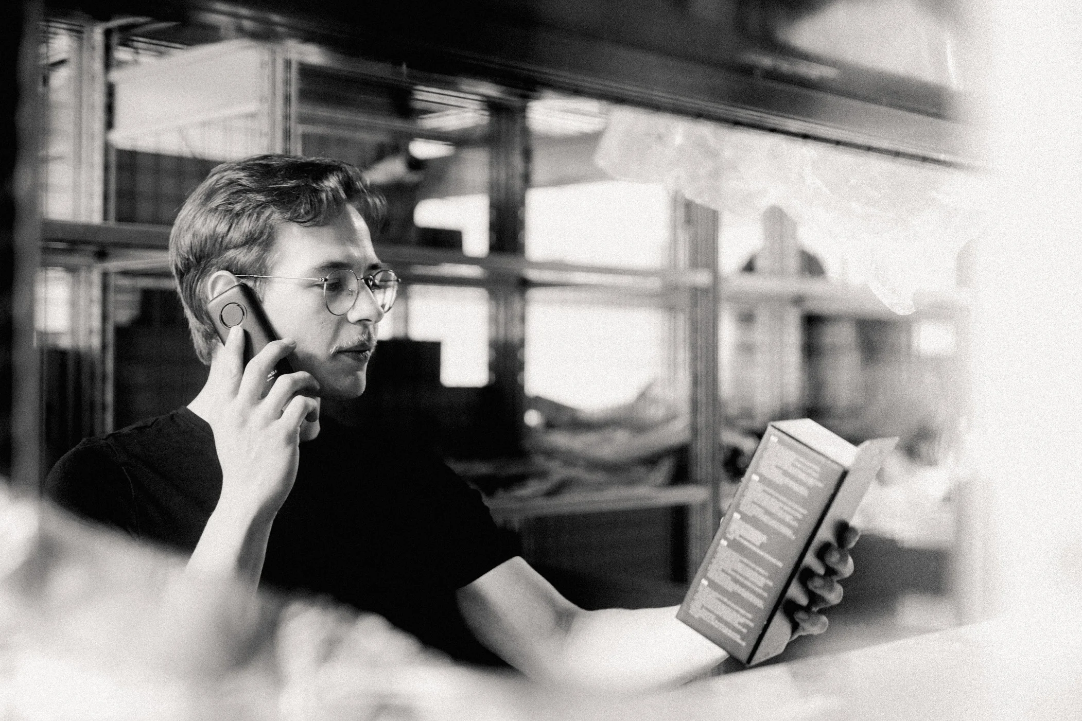 Ein Teiledienstmitarbeiter steht im Lager und trägt ein schwarzes AVEMO T-Shirt. Mit der rechten Hand hält er sich ein Telefon ans Ohr und telefoniert. In der linken Hand hält er eine Produktverpackung vor sich.