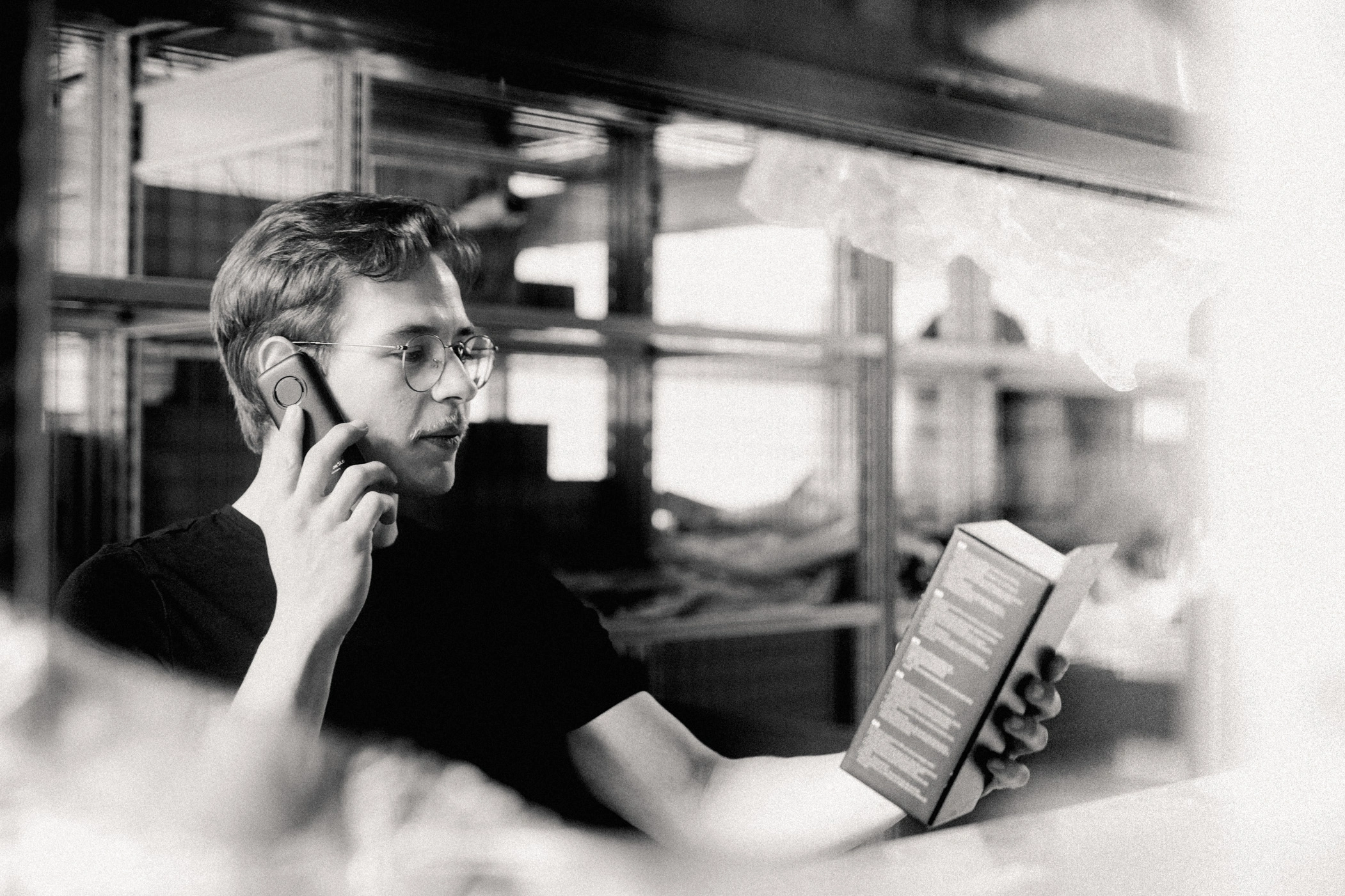 Teiledienst Telefonat m Ein Teiledienstmitarbeiter steht im Lager und trägt ein schwarzes AVEMO T-Shirt. Mit der rechten Hand hält er sich ein Telefon ans Ohr und telefoniert. In der linken Hand hält er eine Produktverpackung vor sich.