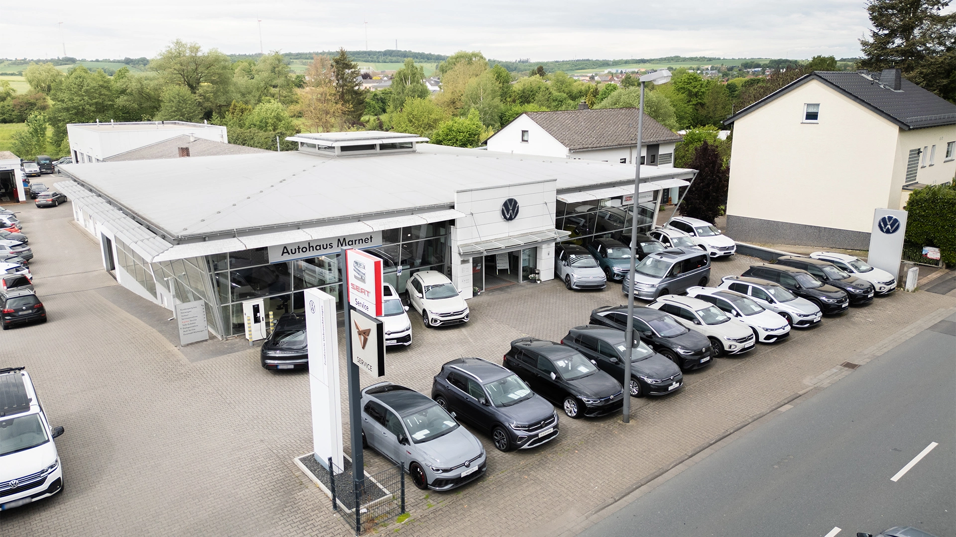 Volkswagen Bad Camberg Header Eine Aufnahme des Volkswagen Autohauses in Bad Camberg, dass das Gebäude und Volkswagen Fahrzeuge vor dem Gebäude, sowie die Einfahrt zeigt.