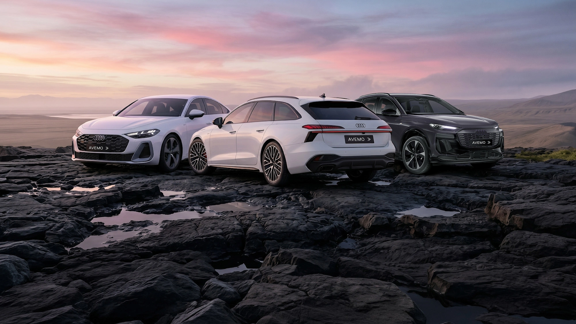 Drei moderne Audi-Modelle – eine weiße Limousine, ein weißer Kombi und ein dunkler SUV – mit AVEMO-Kennzeichen parken auf dunklem, felsigem Terrain. Im Hintergrund erstreckt sich eine weite Landschaft unter einem farbenfrohen Abendhimmel.