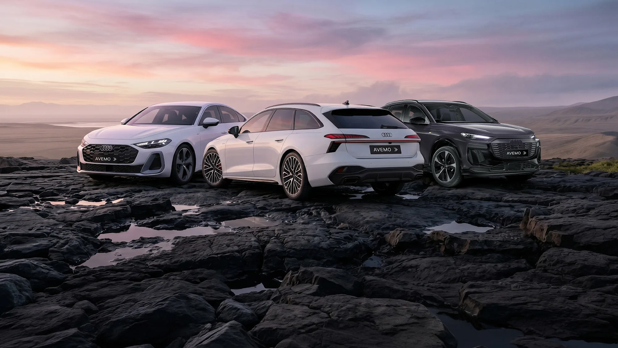 26-040 website Drei moderne Audi-Modelle – eine weiße Limousine, ein weißer Kombi und ein dunkler SUV – mit AVEMO-Kennzeichen parken auf dunklem, felsigem Terrain. Im Hintergrund erstreckt sich eine weite Landschaft unter einem farbenfrohen Abendhimmel.