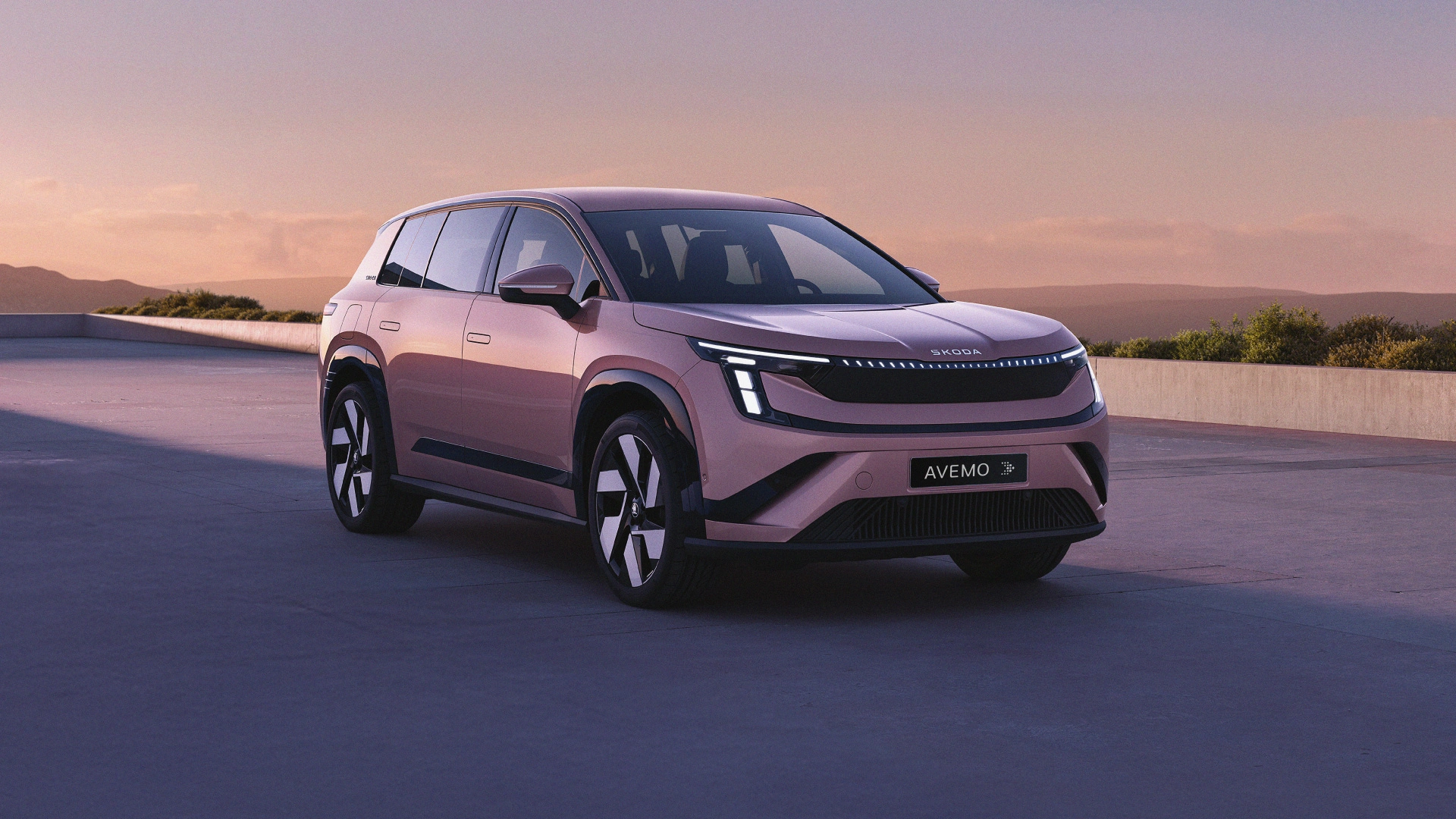 A25-0178 website header Ein moderner, rosé-metallicfarbener Škoda Elroq SUV steht bei Sonnenuntergang auf einer Terrasse mit Pool. Das Auto zeigt markante LED-Scheinwerfer und das Kennzeichen „AVEMO“. Im Hintergrund sind Hügel unter einem sanften, abendlichen Himmel zu sehen.