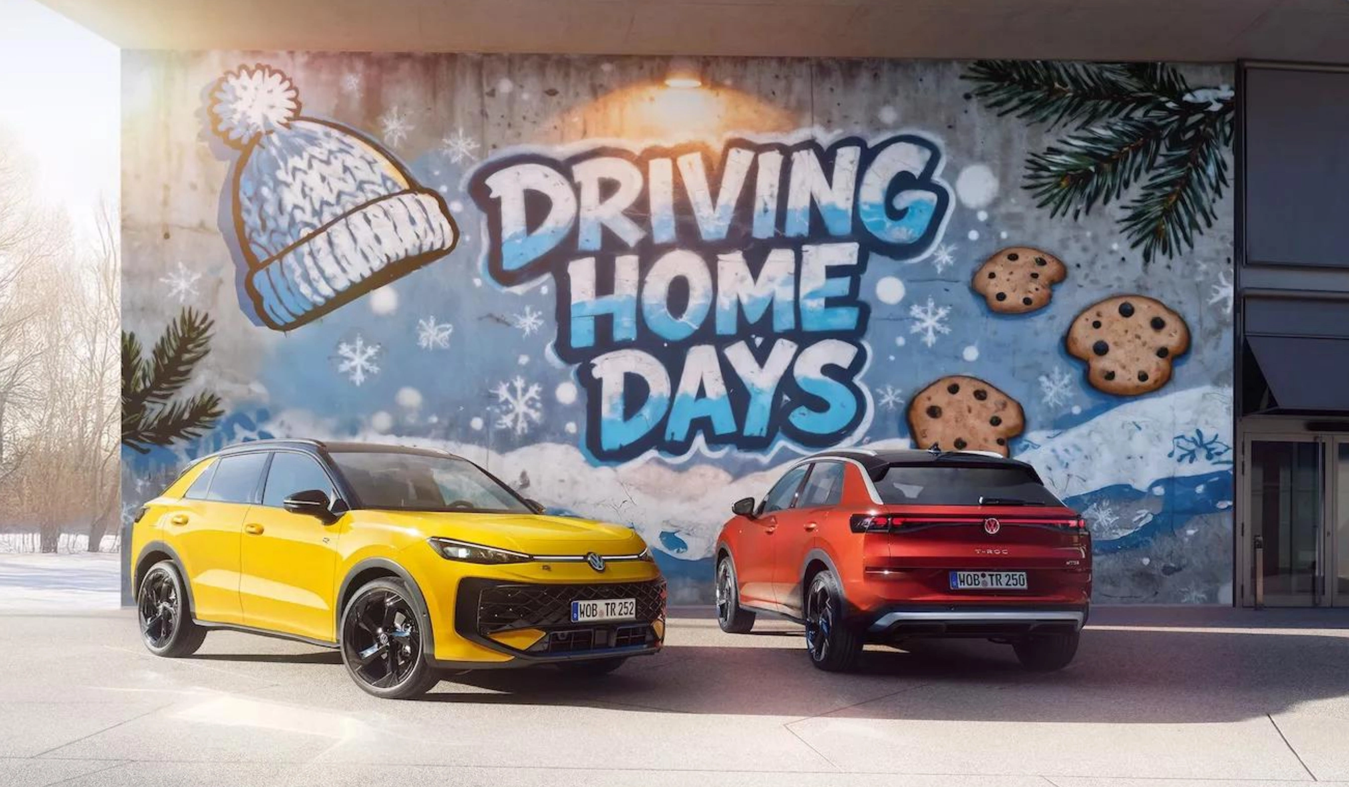 Zwei VW-Fahrzeuge, ein gelber und ein roter SUV, stehen vor einer Mauer mit Graffiti. Das Graffiti zeigt eine Mütze, Schneeflocken, Plätzchen und den Schriftzug "DRIVING HOME DAYS". Das Bild hat einen winterlichen, sonnigen Hintergrund.