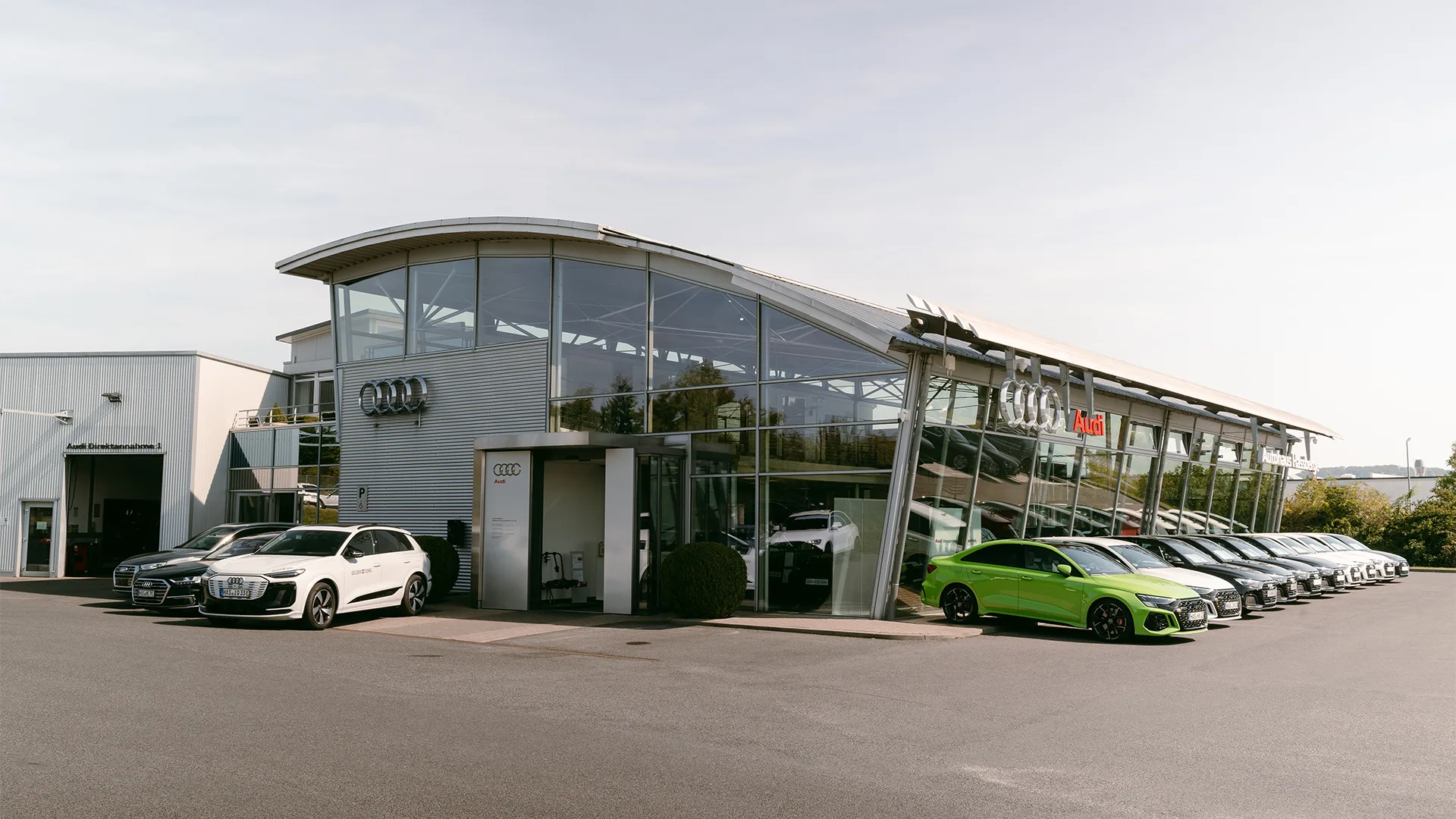 Eine Aufnahme des Audi Autohauses in Haßfurt, dass das Gebäude und Audi Fahrzeuge vor dem Gebäude sowie die Einfahrt zeigt.