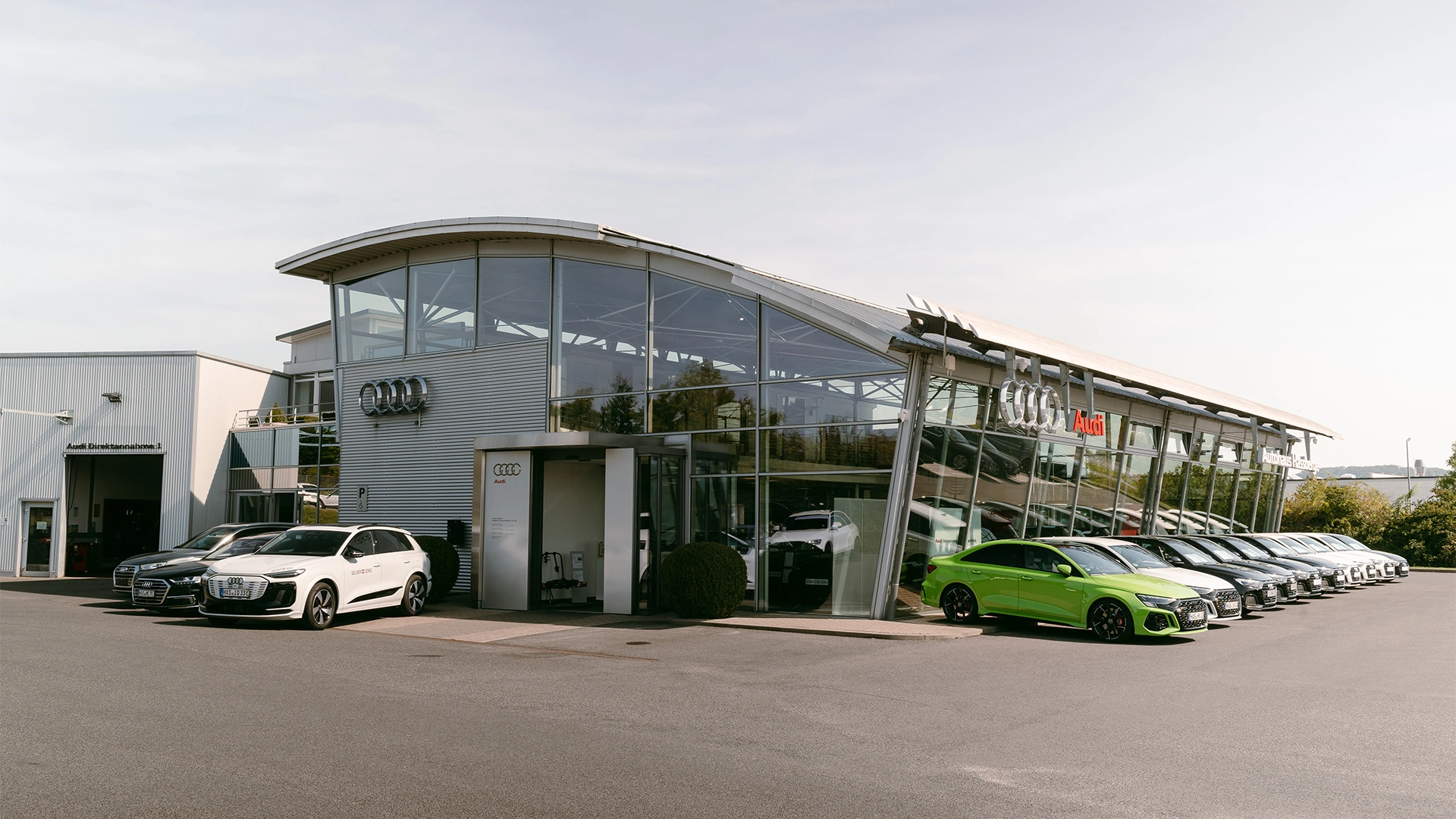 Eine Aufnahme des Audi Autohauses in Haßfurt, dass das Gebäude und Audi Fahrzeuge vor dem Gebäude sowie die Einfahrt zeigt.