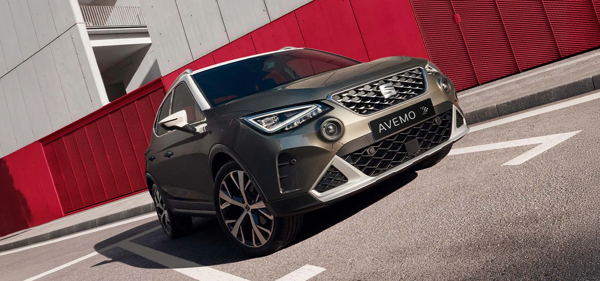 A25-0143 AVEMO header Ein grauer CUPRA Arona mit weißem Dach und schwarzer AVEMO-Plakette im Kühlergrill parkt schräg auf einer Straße. Im Hintergrund sind moderne Gebäude mit roten und weißen Fassaden zu sehen.