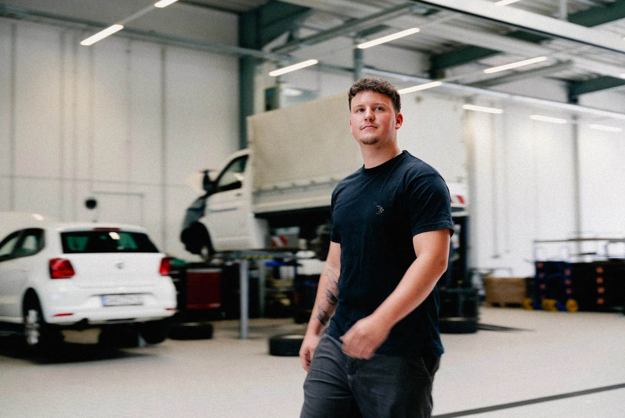 Ein Mann mit schwarzem AVEMO T-Shirt läuft durch die Werkstatt des Autohauses. Im Hintergrund sieht man ein Auto und einen Transporter mit Ladefläche, an denen gearbeitet wird.