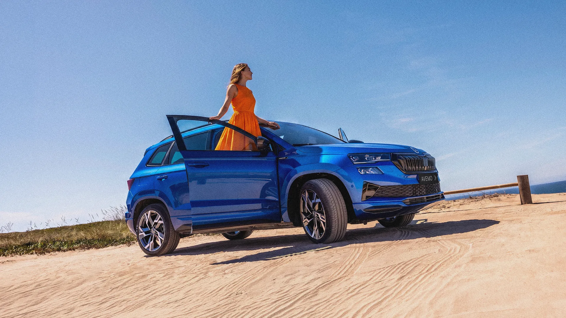 Ein leuchtend blauer Skoda SUV steht mit geöffneter Fahrtür auf einer sandigen Düne vor strahlend blauem Himmel. Eine Frau in einem auffälligen orangen Kleid steht an der geöffneten Tür des Fahrzeugs und blickt in die Ferne.