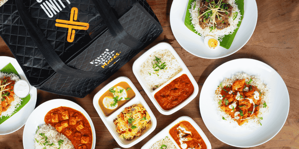 CookUnity insulated delivery bag surrounded by chef-prepared plant-based meals including paneer curry, biryani, and tikka masala served in white containers and plates