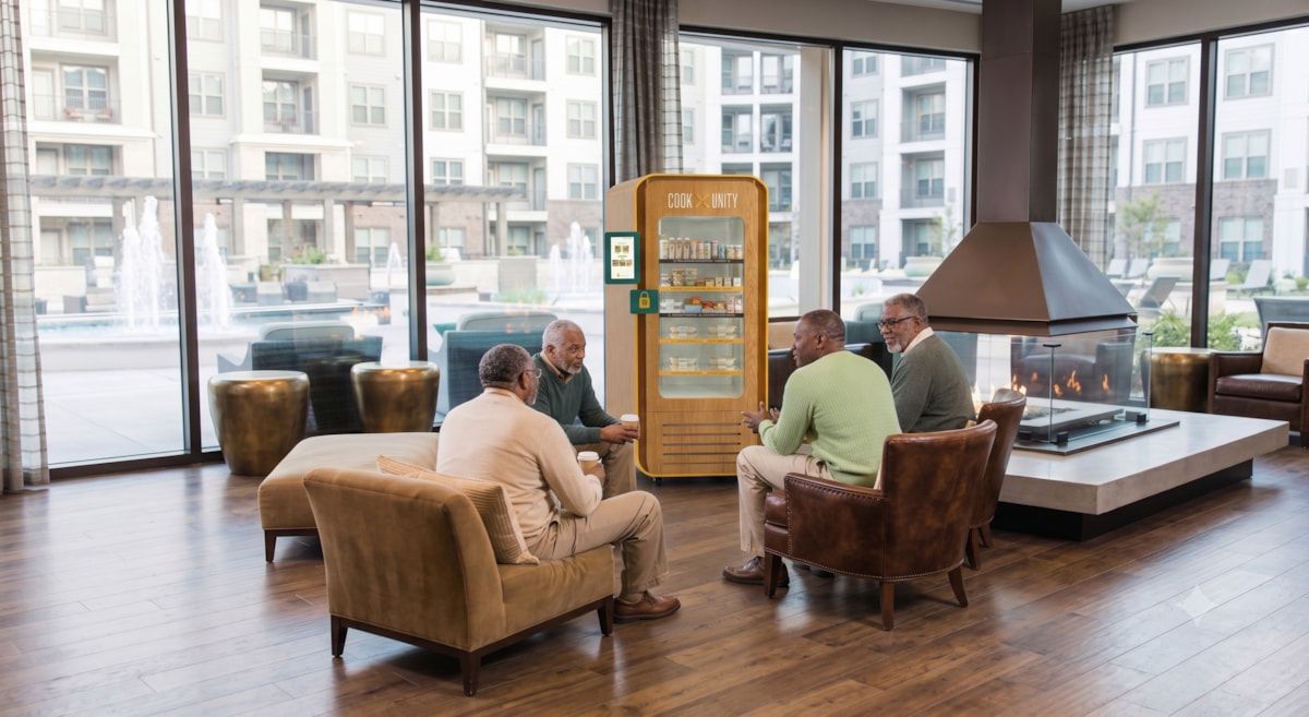 Four residents socializing in a luxury apartment building lounge next to a CookUnity smart fridge with no kitchen or staff required