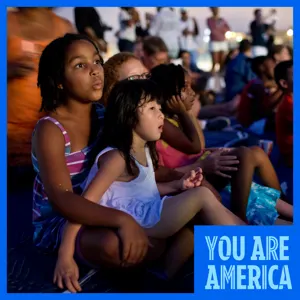 Children sit together outdoors at dusk, watching a performance with focused expressions as a crowd gathers behind them.