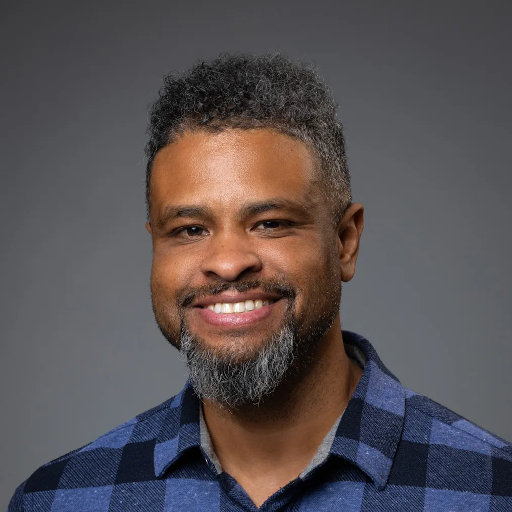 A smiling man with medium-brown skin, short hair and a goatee wearing a blue plaid shirt