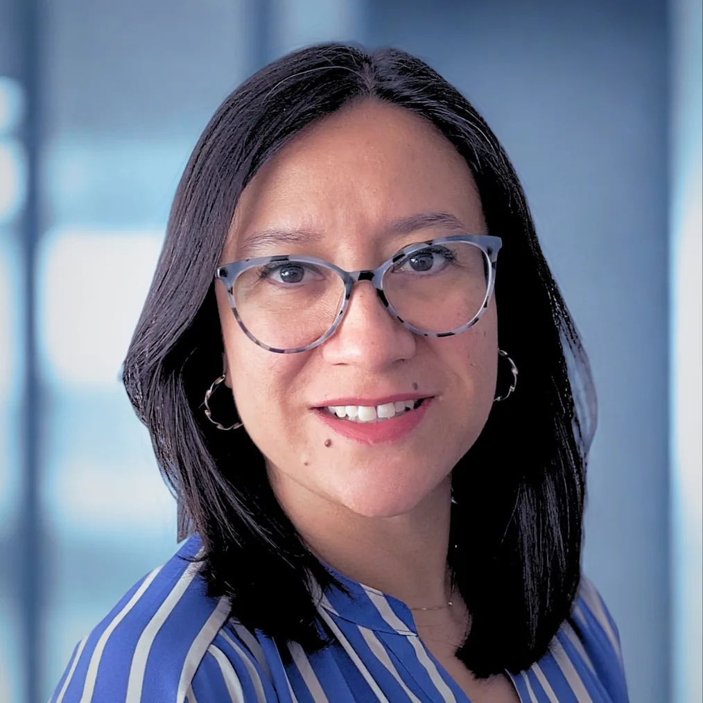 A headshot of Nancy Villafranca Guzman, a person with a medium-light skin tone and straight, dark shoulder-length hair. Facing forward with a gentle smile, she wears patterned, clear-framed glasses and hoop earrings. She is dressed in a blue and white striped blouse against a blurred, light blue background.