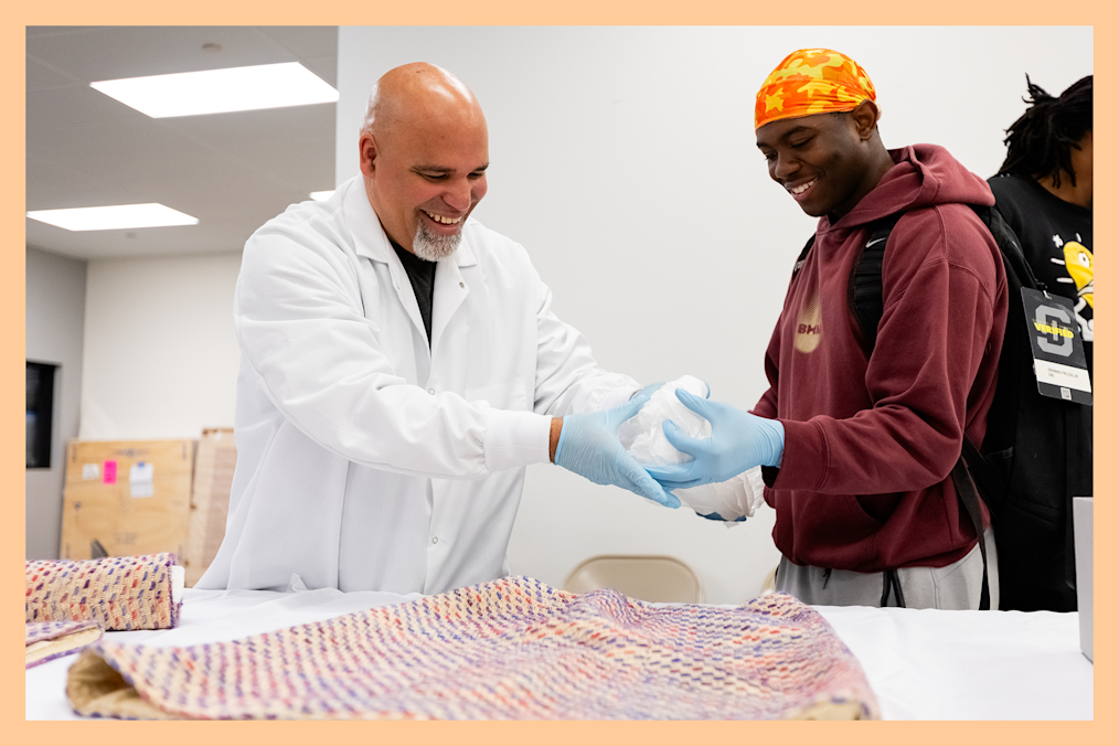 An archivist in a white lab coat and blue gloves shows an archival object to a smiling visitor wearing a maroon hoodie and orange durag.