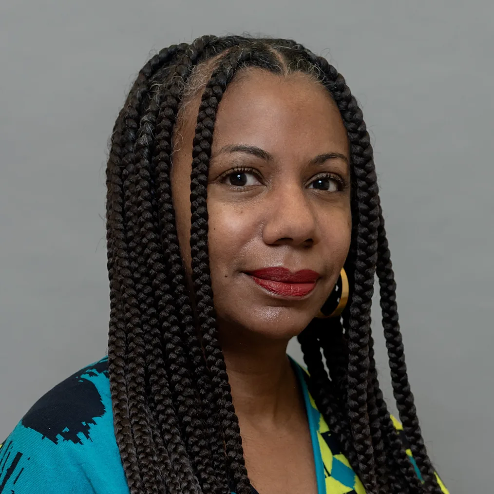 A headshot of Natalie Moore, a Black woman with a medium-dark skin tone and long, dark box braids parted down the middle. Looking forward with a subtle smile and red lipstick, she wears a black, teal, and lime green patterned top. 
