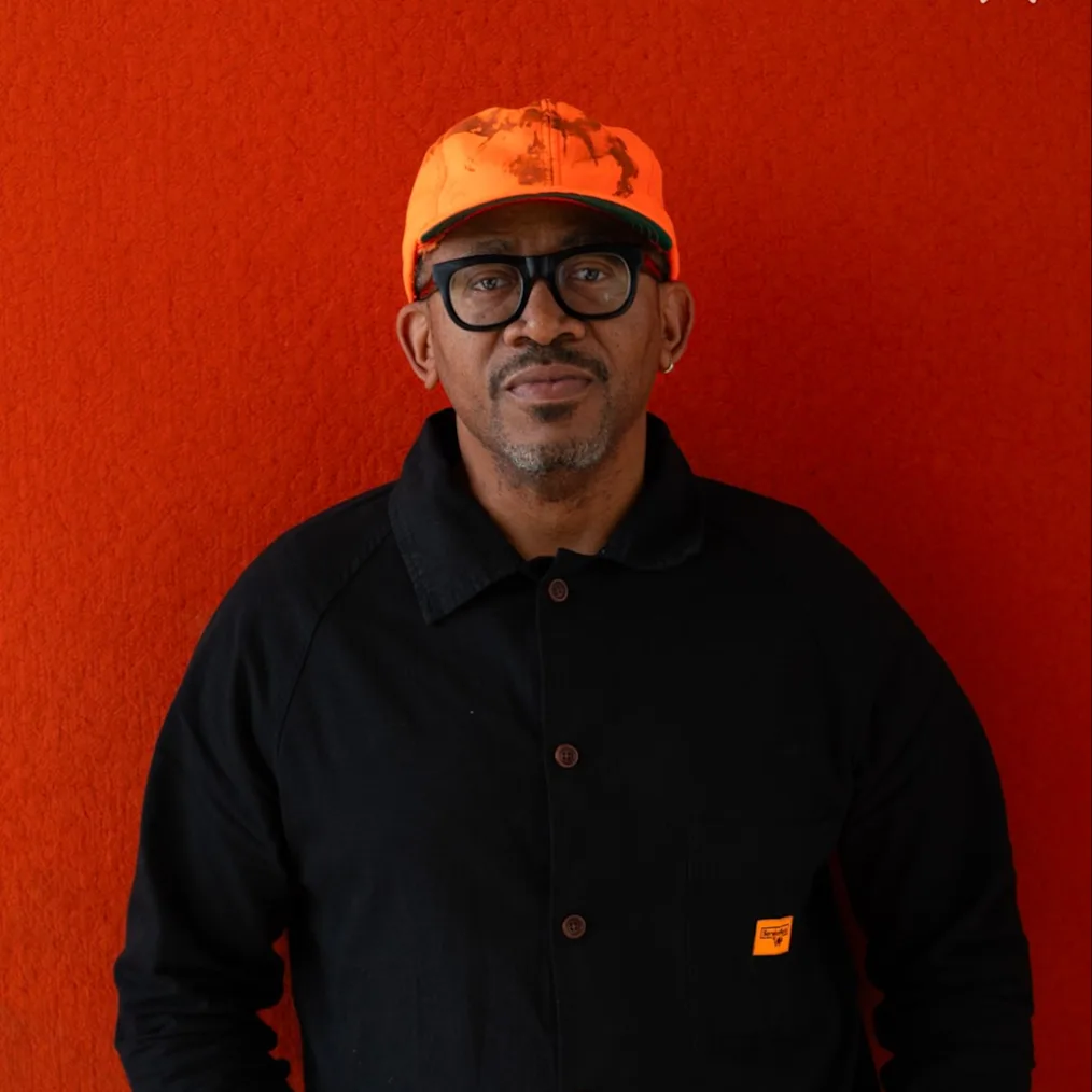 A portrait of Norman Teague, a Black man with a medium-dark skin tone, a mustache, and a graying goatee. He stands facing forward with his hands in his pockets, wearing thick-rimmed black glasses, a bright orange cap, and a black button-down jacket over dark jeans. He stands against a vibrant, textured red background.