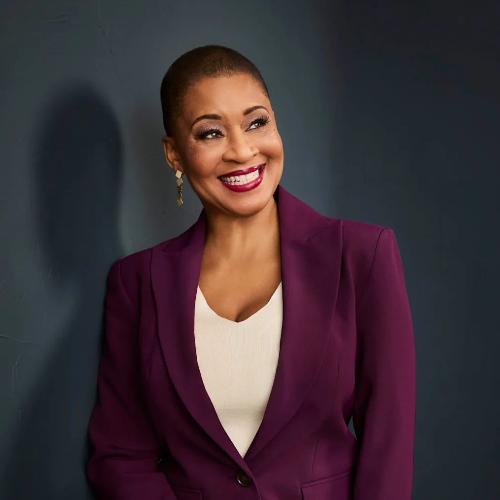 A vertical studio portrait of Jacqueline Stewart, a Black woman with a medium skin tone and closely cropped hair, smiling warmly as she looks off-camera to the right. She wears a rich purple blazer over a cream-colored top, gold geometric earrings, and vibrant red nail polish. She leans against a textured dark blue wall.
