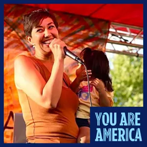 A person with short brown hair and sunglasses holding a young child grabs a mic on an outdoor stage
