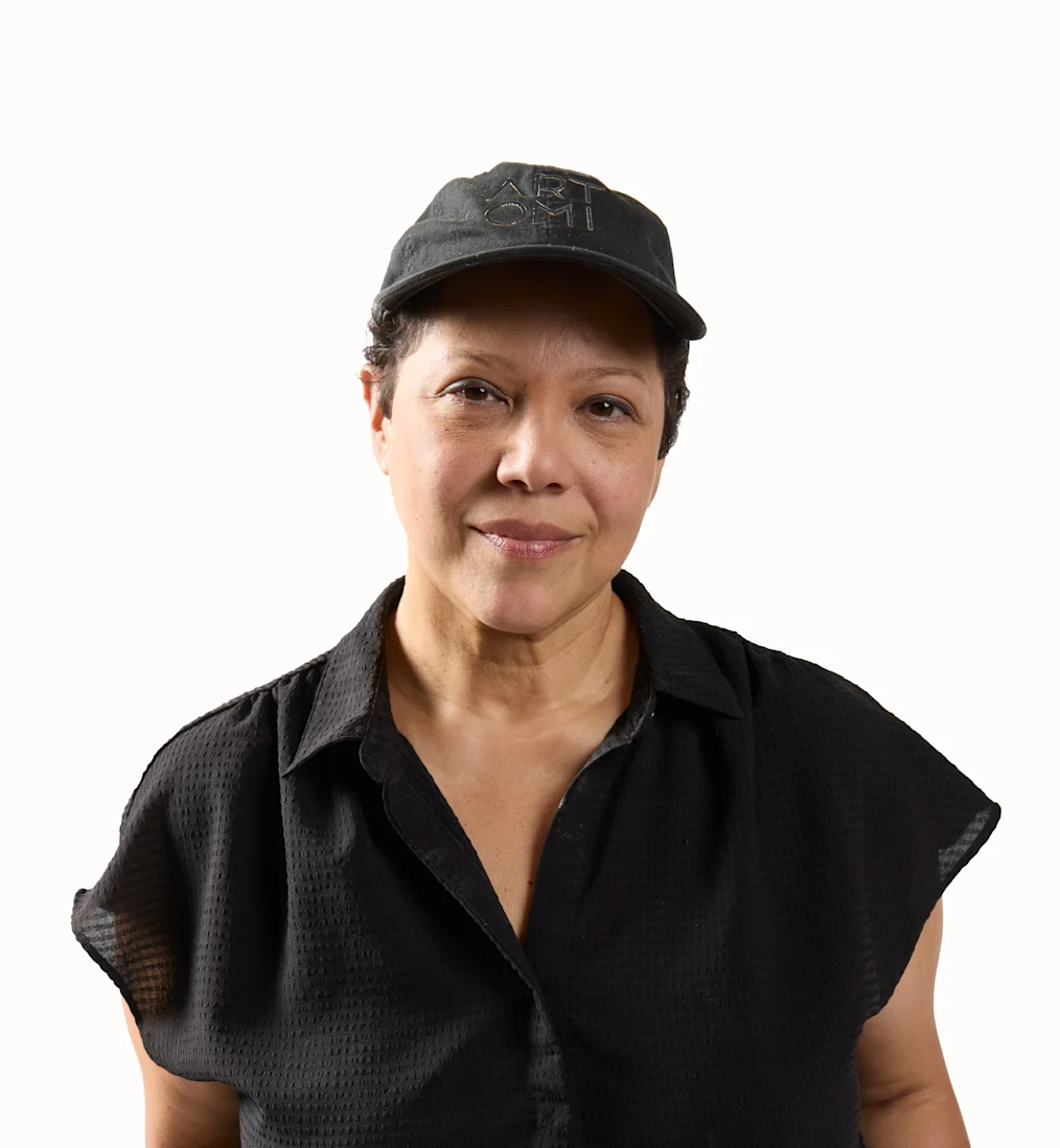 A studio portrait of Edra Soto, a person with a medium-light skin tone and dark hair. Facing forward with a slight, gentle smile, they wear a dark, short-sleeved button-down shirt and a black baseball cap with subtle embroidered lettering 