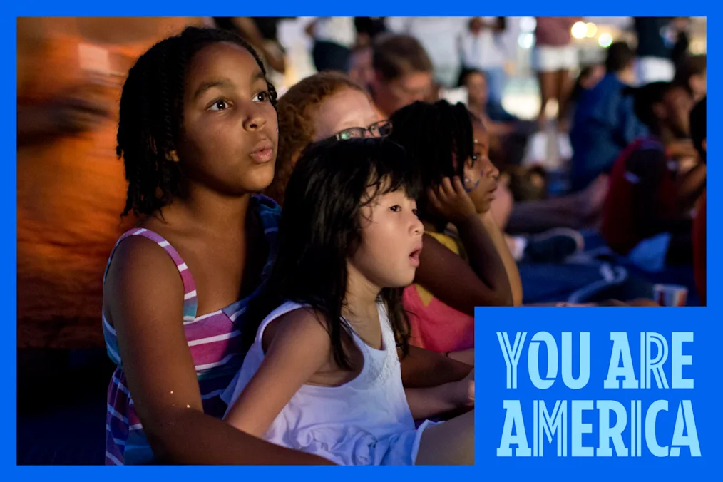 Children sit together outdoors at dusk, watching a performance with focused expressions as a crowd gathers behind them.
