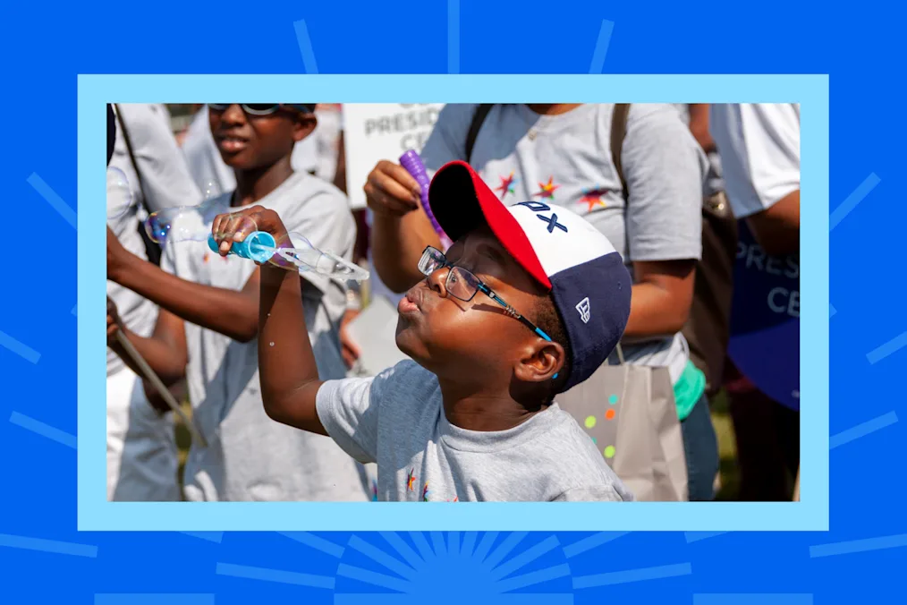 A young boy wearing glasses and a baseball cap blows bubbles at an outdoor community event, framed by a blue graphic border.