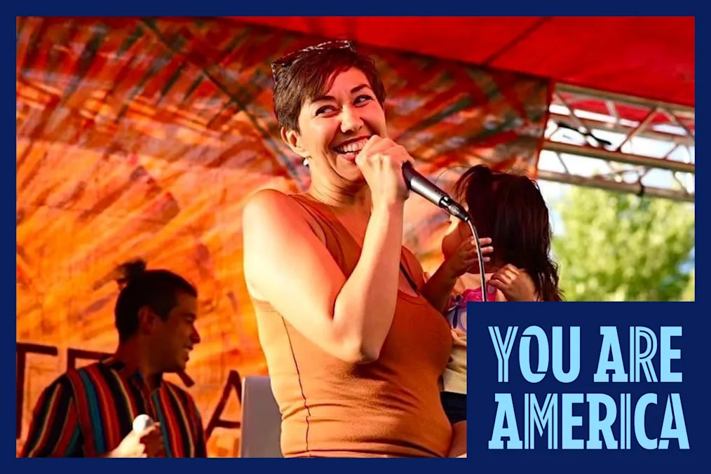 A person with short brown hair and sunglasses holding a young child grabs a mic on an outdoor stage