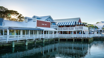 A picturesque Hilton Head waterfront scene at sunset, bursting with warm light and vibrant activity, evokes the lively, festive spirit of a local gathering. The charming coastal buildings on stilts, boardwalk ambiance, and people enjoying the area closely echo the welcoming atmosphere of the Hilton Head Island Shrimp Festival.