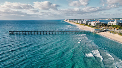 An aerial view of Palm Beach, FL, featuring a long pier stretching into turquoise waters, bordered by a sandy beach and lush greenery, perfectly encapsulates the allure of private beach access and coastal luxury living.