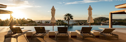 Outdoor pool area facing a panoramic view of the ocean with the sunsetting casting a golden glow over the poolside lounge chairs.