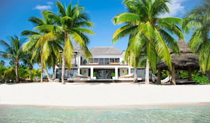 Rear exterior of the home with the private beach leading up to the ocean and lush palm trees placed around the beach with lounge chairs placed underneath them.