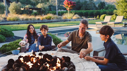 A family of two adults and two children roast marshmallows around a pool-side fire pit. 