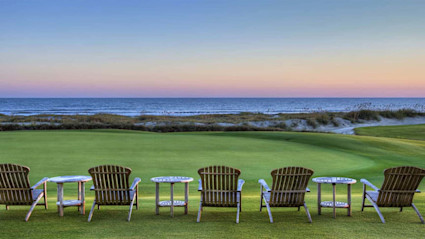This image captures a serene winter sunset on Kiawah Island, SC, with wooden Adirondack chairs arranged on a manicured lawn overlooking sand dunes and the ocean. The soft pastel sky and tranquil setting evoke a sense of peaceful seclusion and luxury, perfectly representing the exclusive winter retreat experience offered on Kiawah Island.