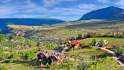 A scenic landscape in Telluride, Colorado, with lush green hills, wooden homes nestled among trees, and majestic mountains in the background. This tranquil mountain setting exudes privacy and natural beauty, perfectly encapsulating the concept of luxury homes offering secluded access to nature.