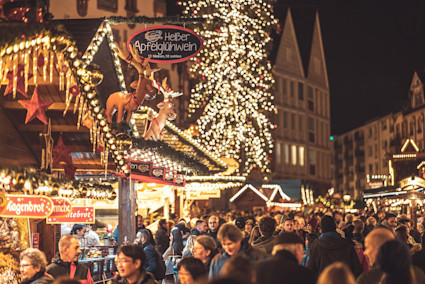 A crowded market square with white lights lining the trees and stalls selling mulled wine and seasonal treats.