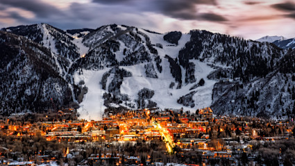 Twilight view of Aspen aglow with warm lights nestled beneath snow-capped mountains, capturing the festive magic and winter wonder of a luxury holiday season in the Colorado Rockies.