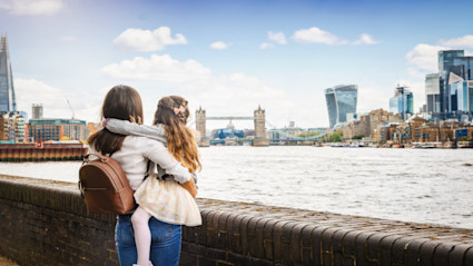 A woman and a young girl stand by the River Thames with Tower Bridge and London's distinctive skyline in the background, capturing the spirit of autumn exploration and family travel in London. The image conveys a sense of connection to the city's culture and iconic sights, evoking the essence of fall adventures.