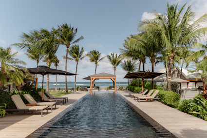 Long lap pool flanked by palm trees and lounge chairs leads toward a wooden gazebo and the ocean in St. Barth.