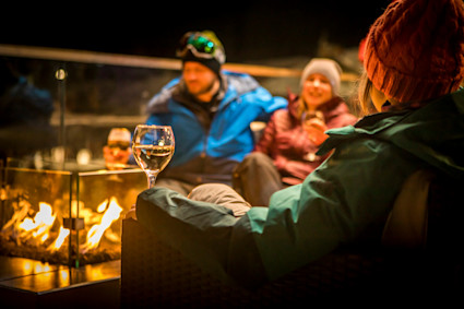 Three people in snow gear gather around a fire at night with glasses of wine.